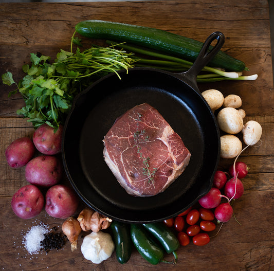 Raw roast in a cast iron skillet surrounded by vegetables on a wooden surface